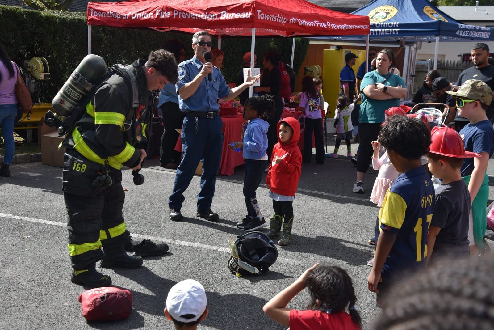 A firefighter shows children the gear and air pack worn on the job while a department member explains.